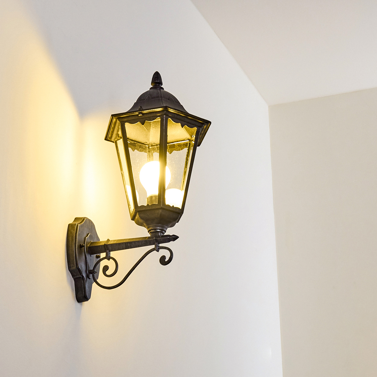 Eine an der Wand montierte, verzierte schwarze Laterne mit einer leuchtenden Glühbirne. Sie befindet sich an einer weißen Wand. Das Licht wirft einen warmen Schein.