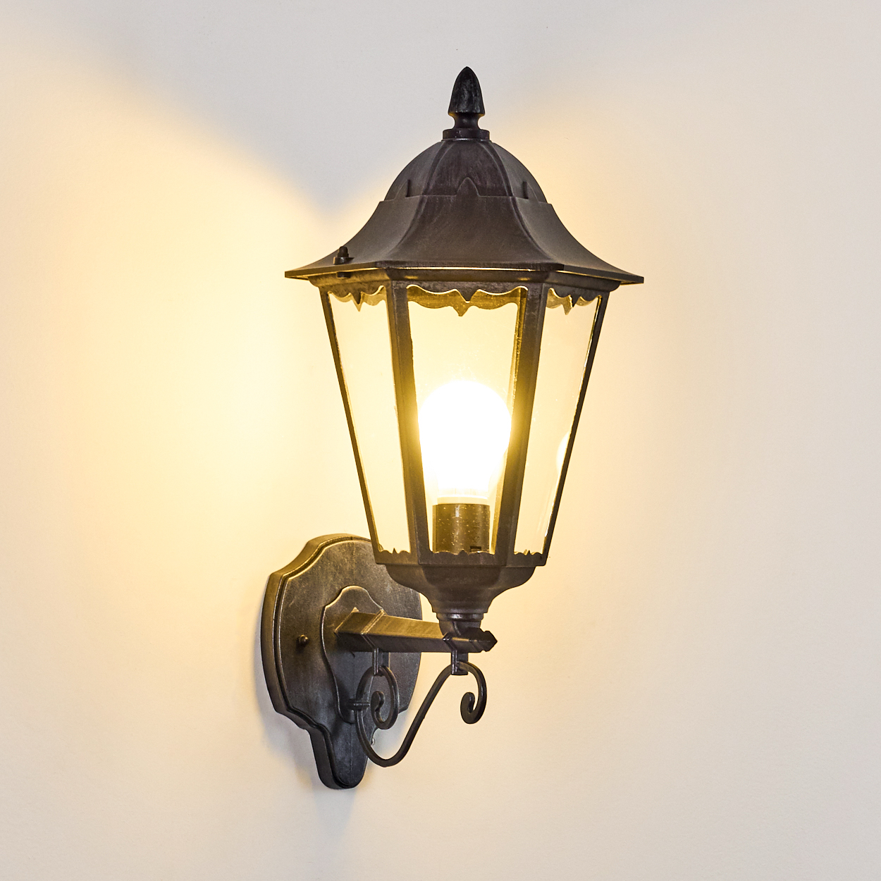 Eine schwarze Außenlampe mit einer brennenden Glühbirne ist an einer beigefarbenen Wand befestigt. Die Lampe hat ein dekoratives Design und Glasscheiben.