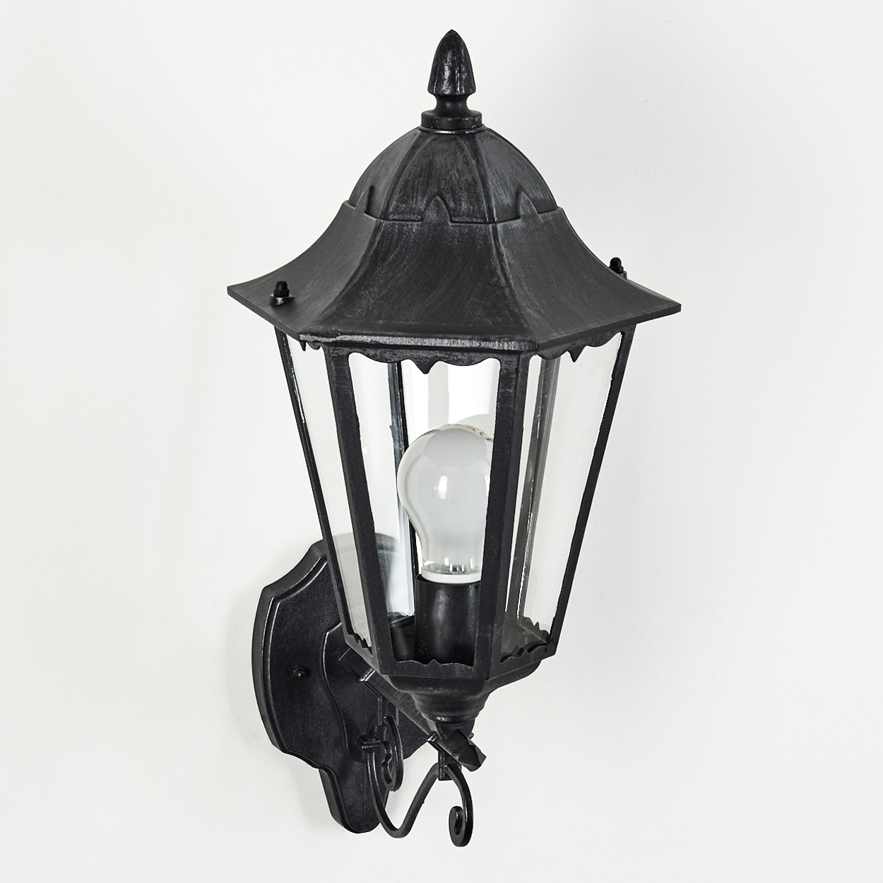 Eine schwarze Außenlampe ist an einer weißen Wand montiert. Die Lampe hat ein Glasgehäuse, eine Glühbirne und dekorative Metallarbeiten.