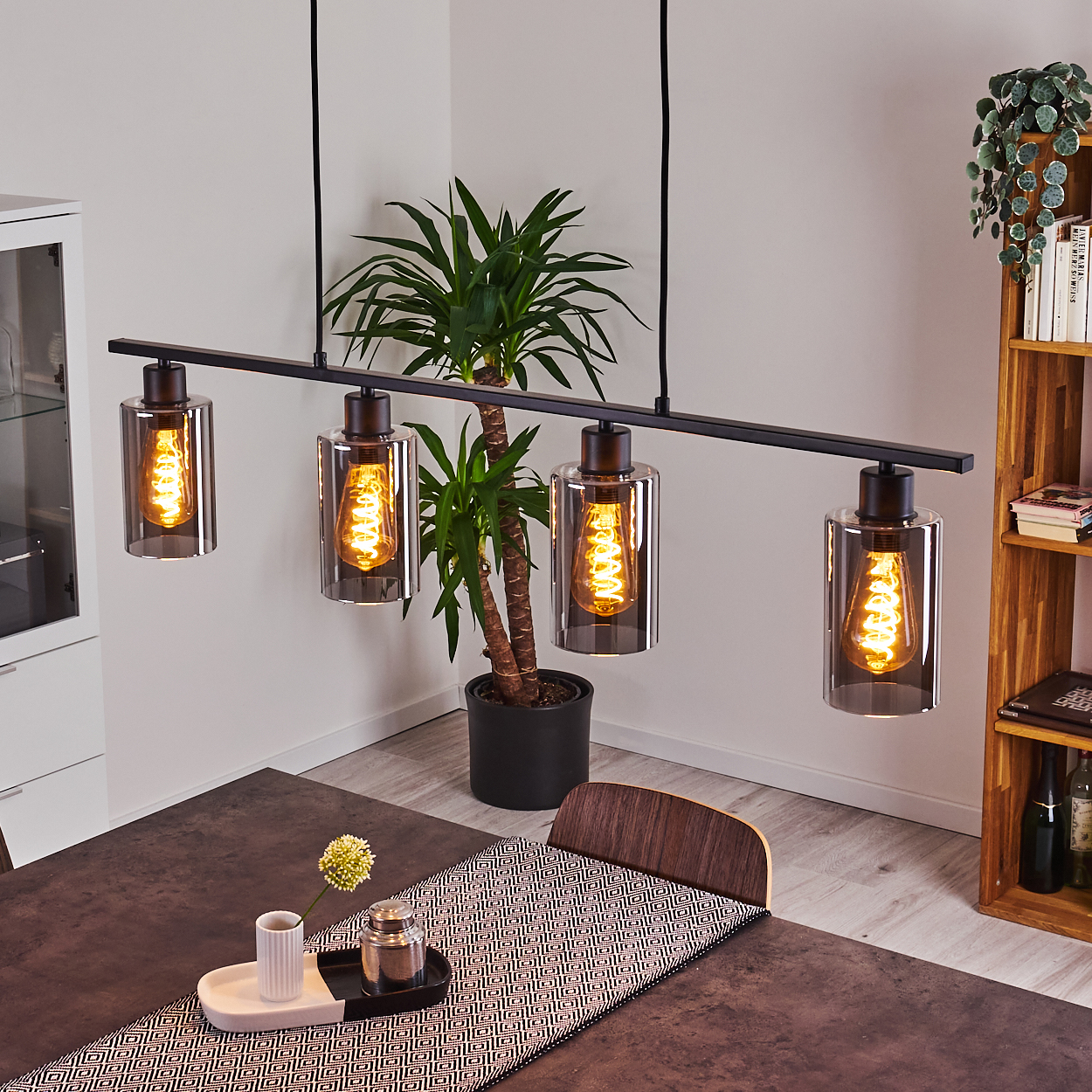 Esszimmer mit schwarzer Leuchte, Tisch, Pflanze und Holzregal mit Büchern und Dekorationen.