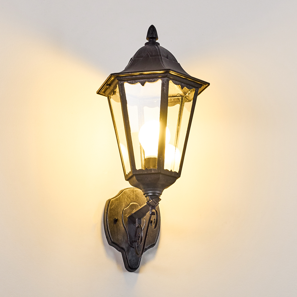 Eine verzierte, schwarze Außenlampe mit einer leuchtenden Glühbirne. Sie ist an einer beigefarbenen Wand montiert.