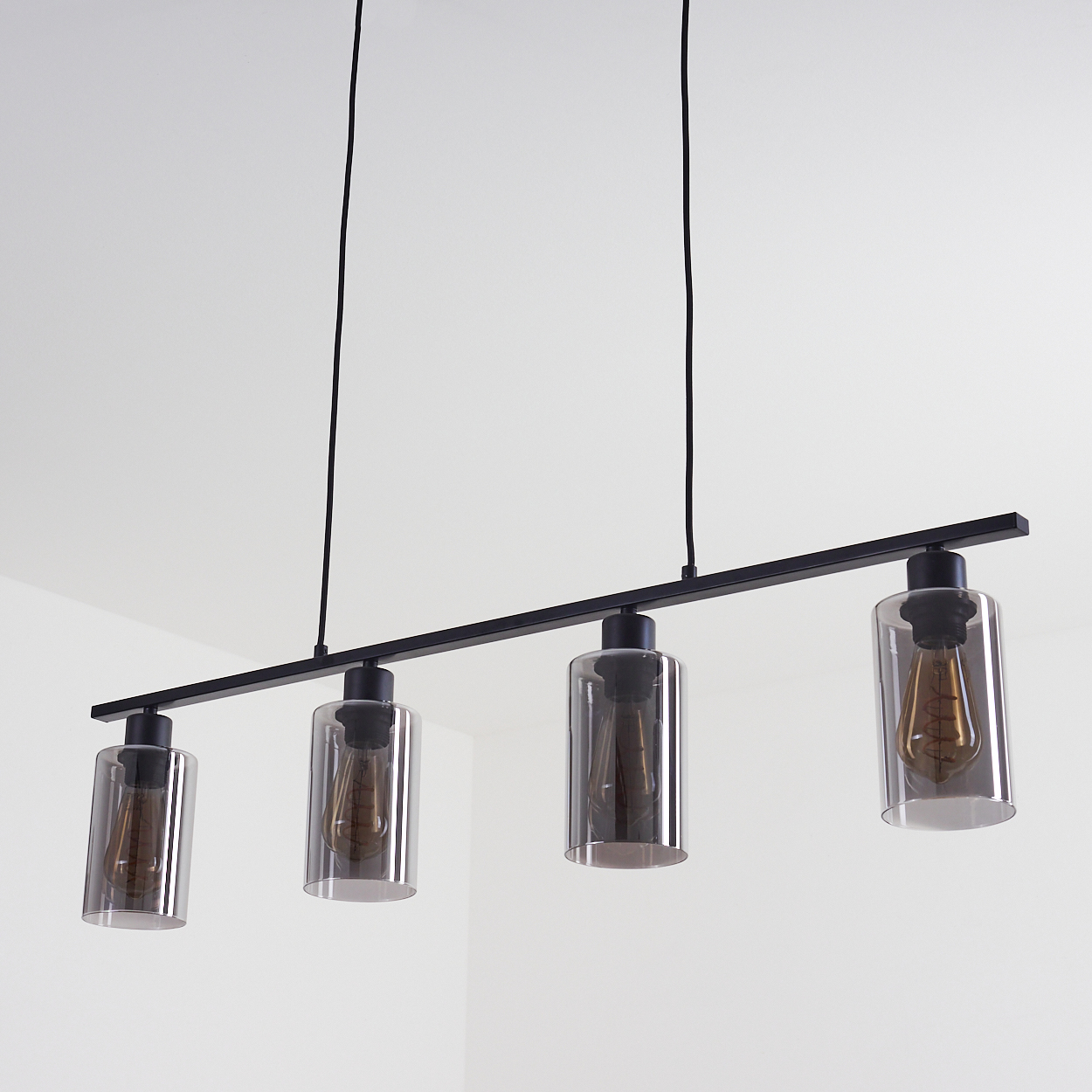 Eine moderne schwarze lineare Hängeleuchte mit vier zylindrischen Glasschirmen, die jeweils eine Glühbirne enthalten, hängt vor einem weißen Hintergrund.