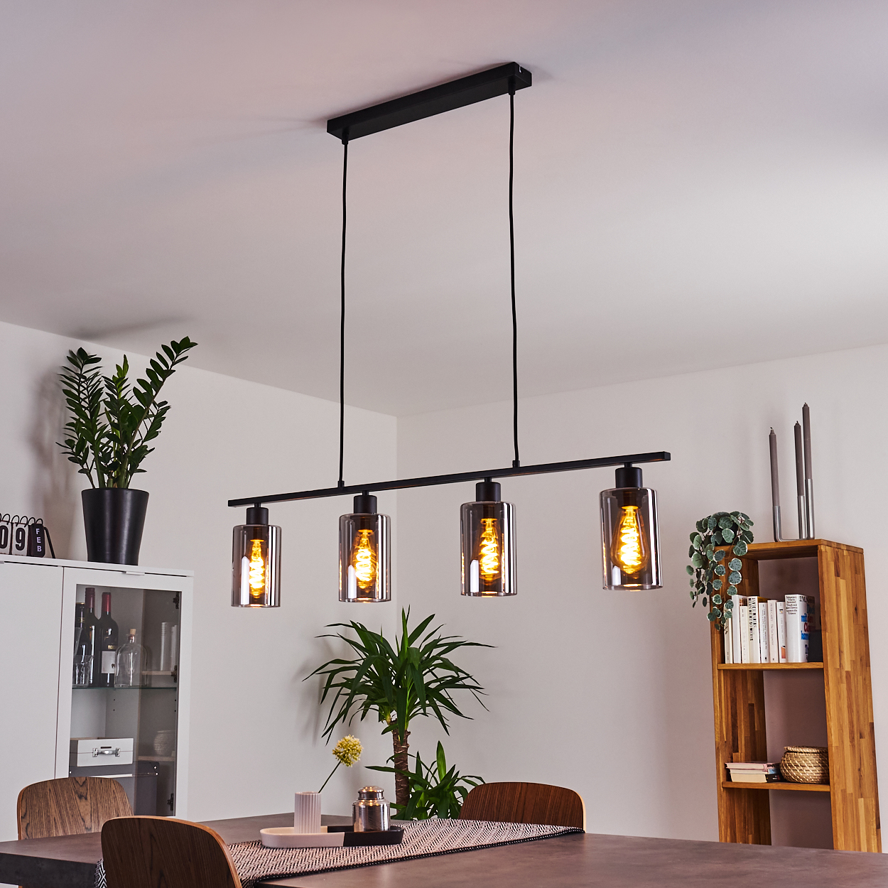 Ein modernes Esszimmer mit einer schwarzen Leuchte mit vier Glühbirnen. Es gibt Pflanzen, einen Schrank, ein Holzregal und einen Esstisch.
