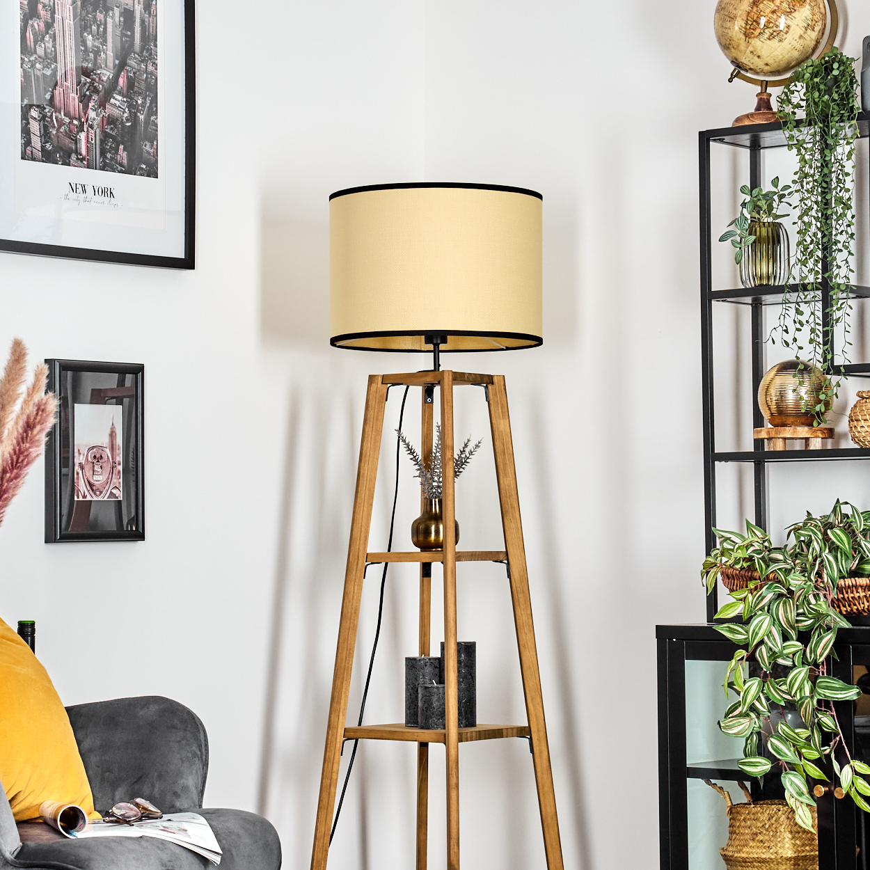 Eine hölzerne Stehlampe mit beigefarbenem Schirm steht in einem Raum. Regal mit Pflanzen und einem Globus. Kunst und ein grauer Stuhl.