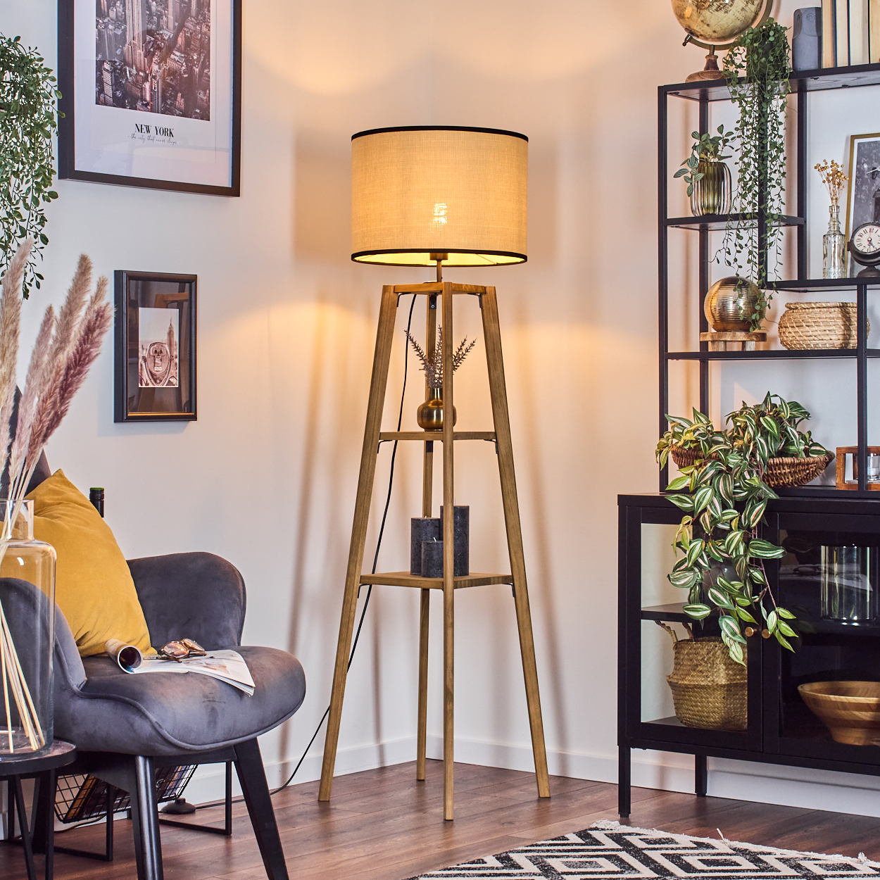 Ein gemütliches Zimmer mit einer Stehlampe aus Holz. Ein grauer Sessel mit einem gelben Kissen und ein schwarzes Regal mit Pflanzen.