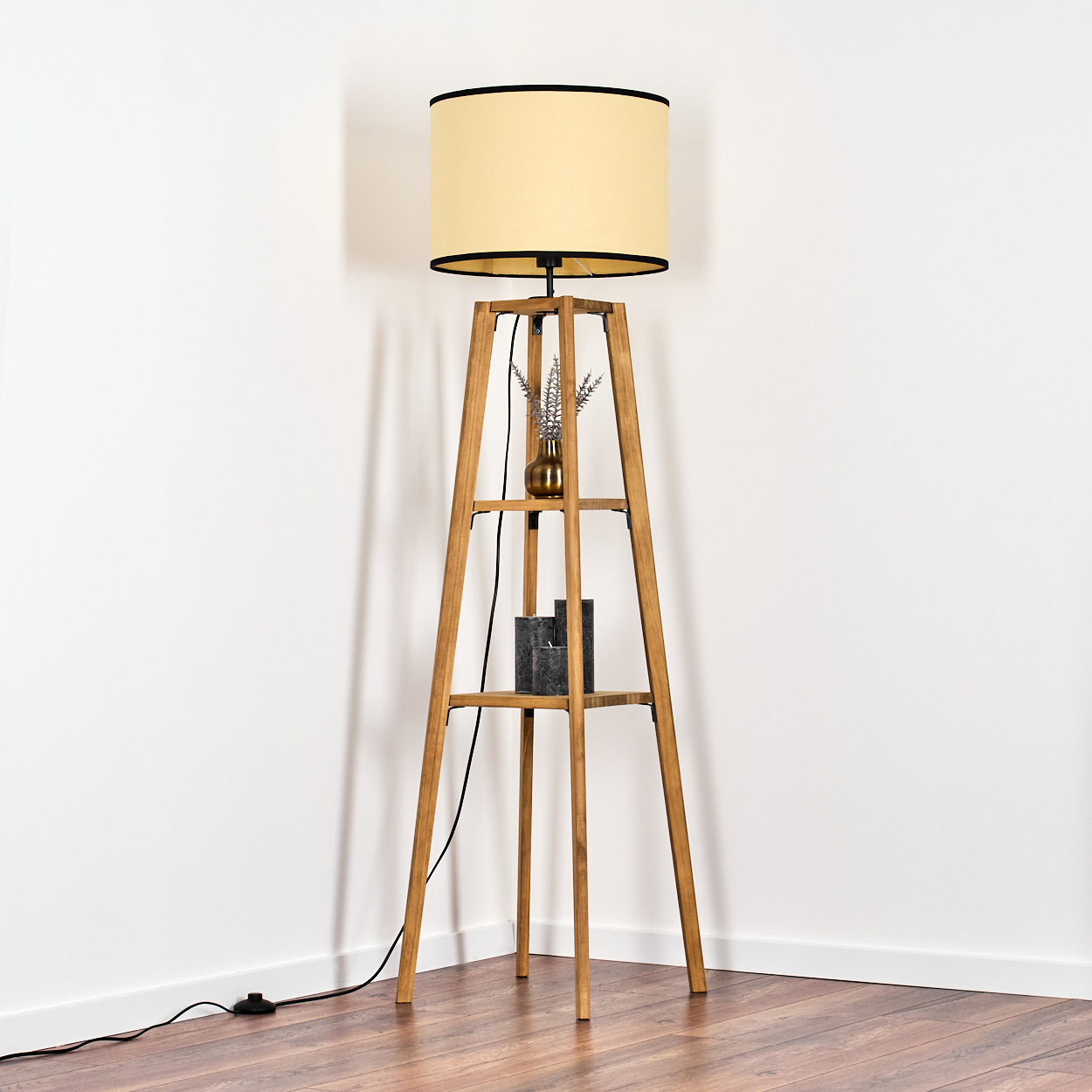Eine Stehlampe aus Holz mit Regalen steht in einer Ecke. Sie hat einen beigen Lampenschirm und ein schwarzes Kabel. Eine Vase und Deko stehen auf den Regalen.