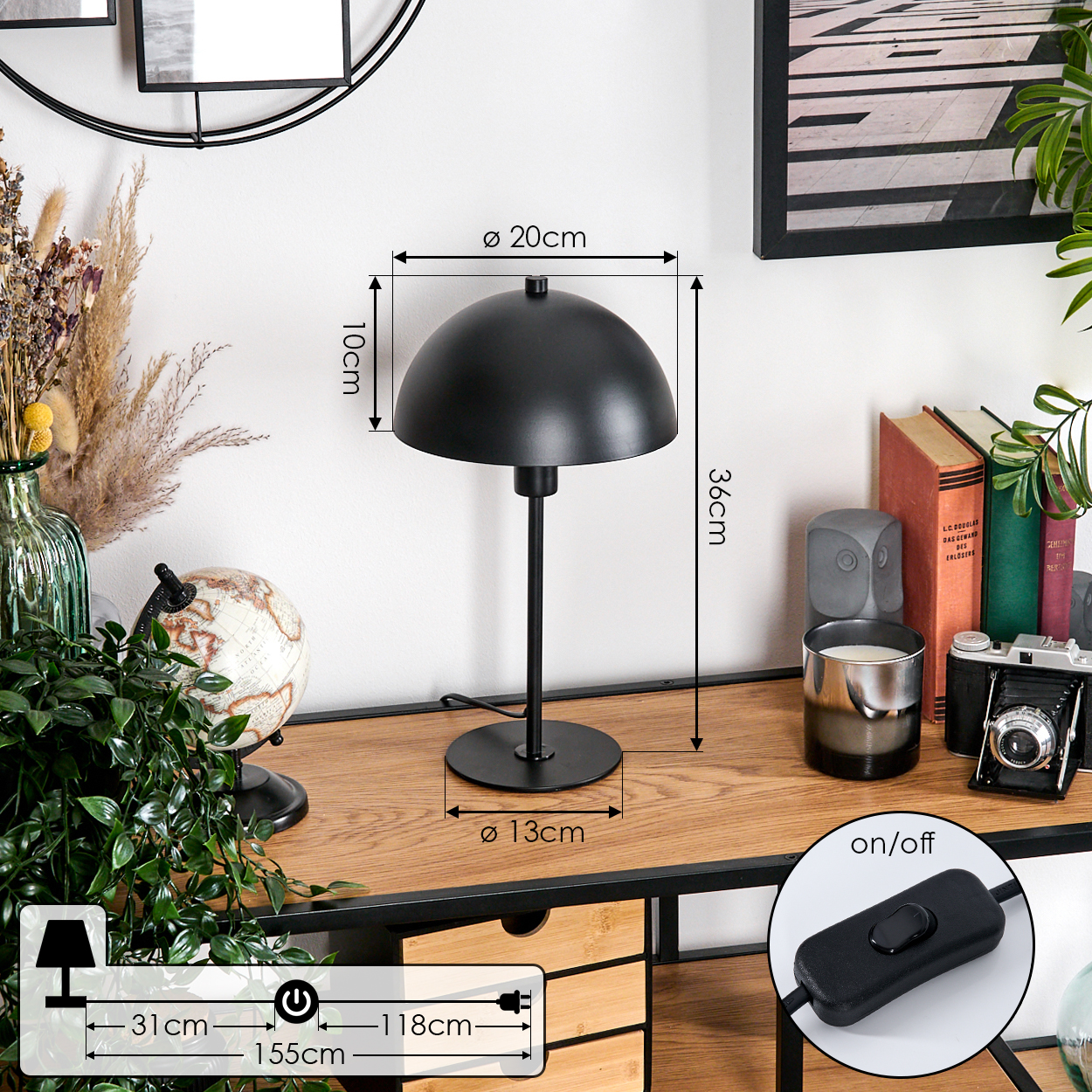 Eine schwarze Schreibtischlampe mit Schirm steht auf einem Holztisch. Bücher, eine Kamera und Pflanzen umgeben sie. Ein/Aus-Schalter ist sichtbar.