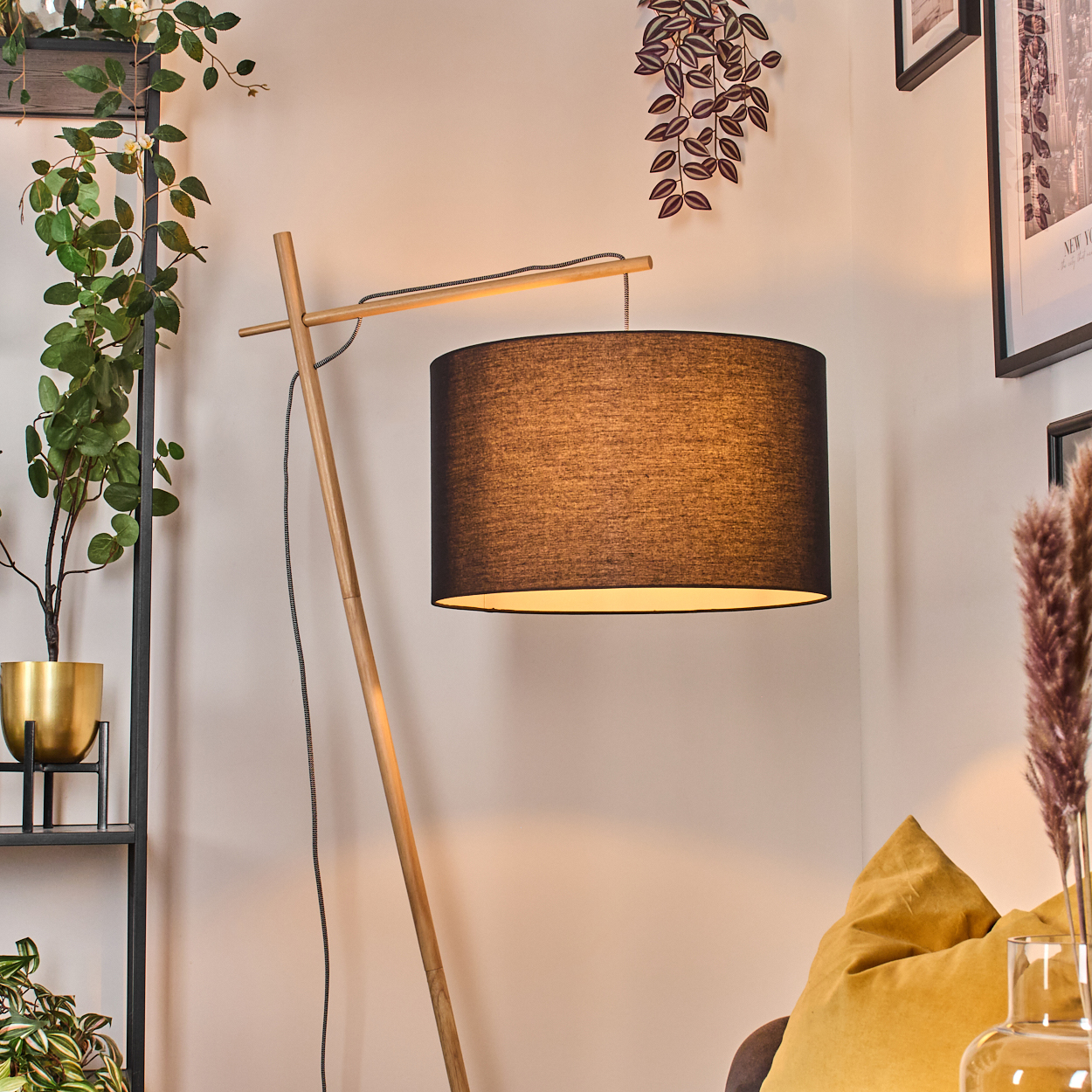 Eine Stehlampe aus Holz mit schwarzem Lampenschirm steht in einer Ecke. Pflanzen und ein goldenes Kissen sind im Hintergrund.