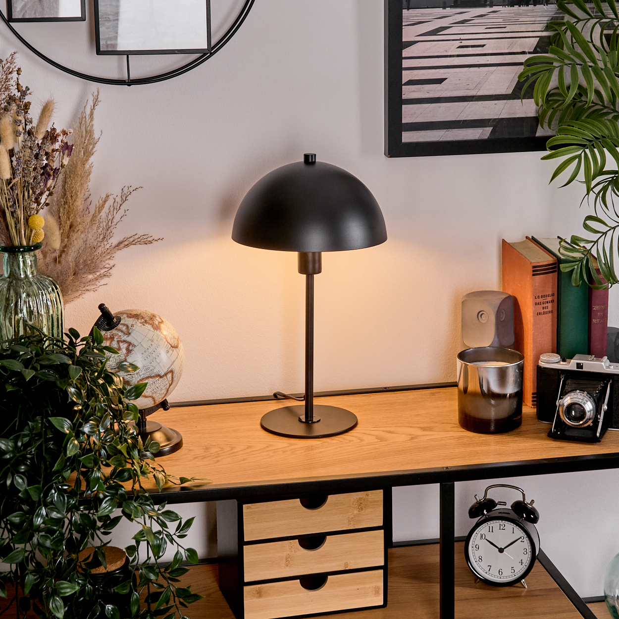 Ein Schreibtisch mit schwarzer Lampe, Büchern, Globus, Kamera und Uhr. Pflanzen und ein gerahmtes Bild sind ebenfalls zu sehen.