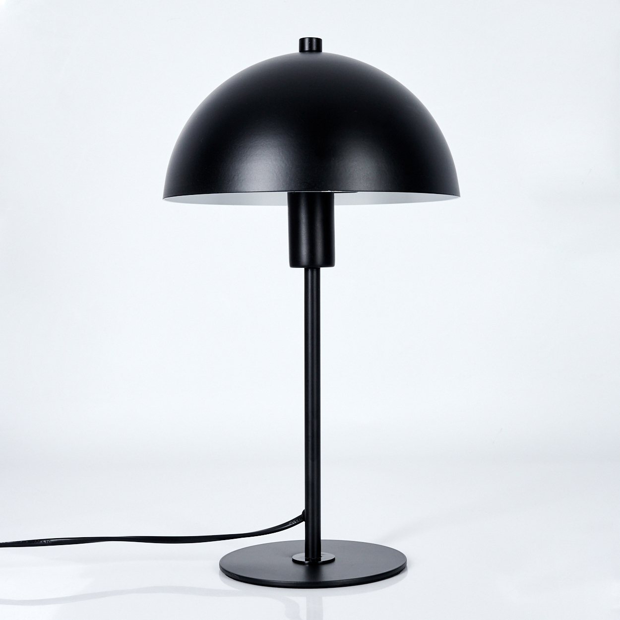 Eine schwarze Tischlampe mit kuppelförmigem Schirm, dünnem Ständer und kreisförmigem Fuß. Der Hintergrund ist weiß. Ein Kabel ist sichtbar.