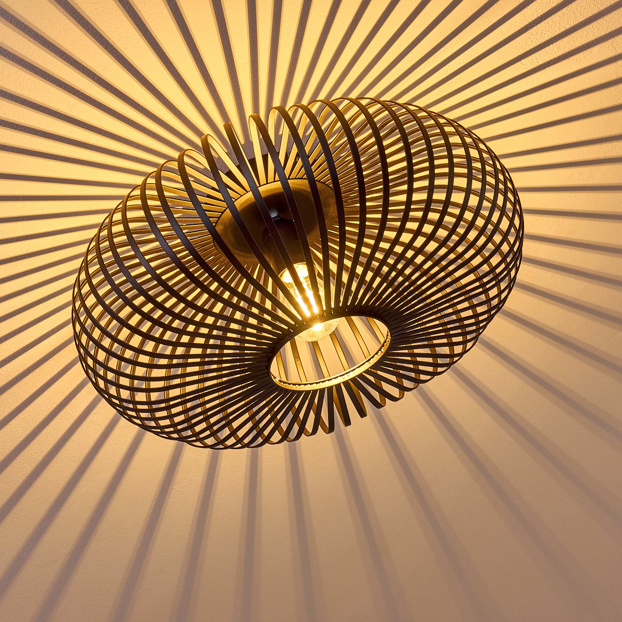 Eine schwarze, kugelförmige, käfigartige Lampe hängt von der Decke. Sie wirft radiale Schatten auf die weiße Wand, die vom Licht der Lampe beleuchtet wird.