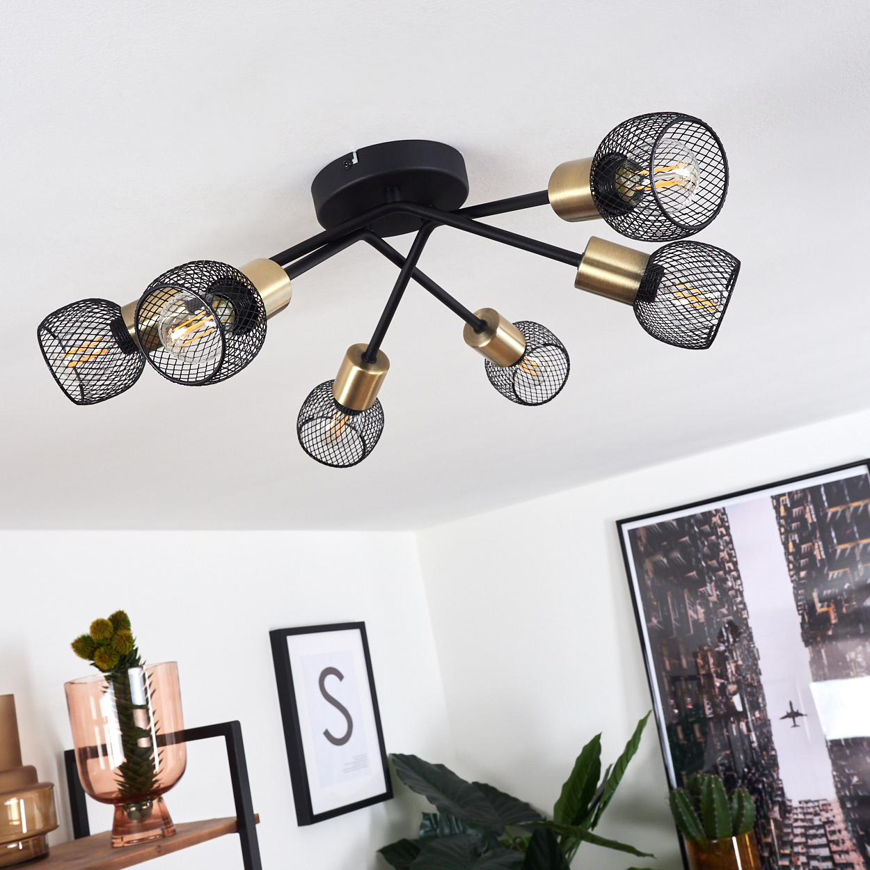 Eine Deckenleuchte mit sechs Lampen. Schwarze Arme mit goldenen Sockeln und Drahtkäfigen. Der Hintergrund hat Dekoration und ein Bild.