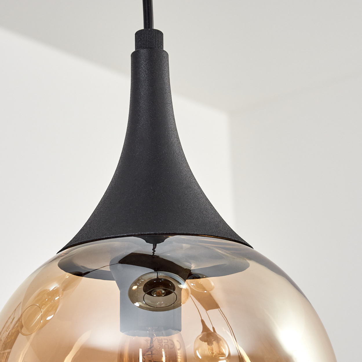 Eine schwarze Pendelleuchte mit bernsteinfarbenem Glasschirm. Die Glühbirne ist sichtbar. Der Hintergrund ist eine weiße Wand.