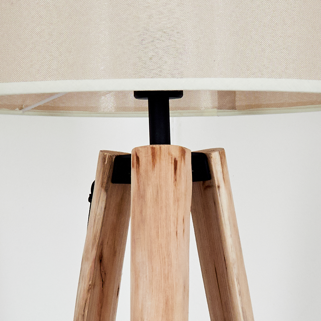 Nahaufnahme einer Holzstativlampe. Beiger Lampenschirm oben. Die Beine sind hellbraunes Holz, schwarze Metallgelenke und ein weißer Hintergrund.