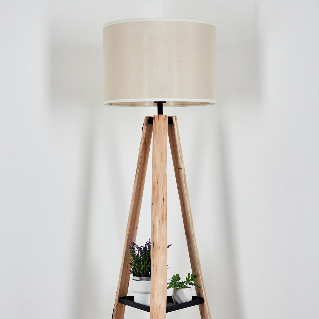 Eine hölzerne Stativlampe mit beigem Lampenschirm steht vor einer weißen Wand. Kleine Topfpflanzen stehen auf einem Regal.