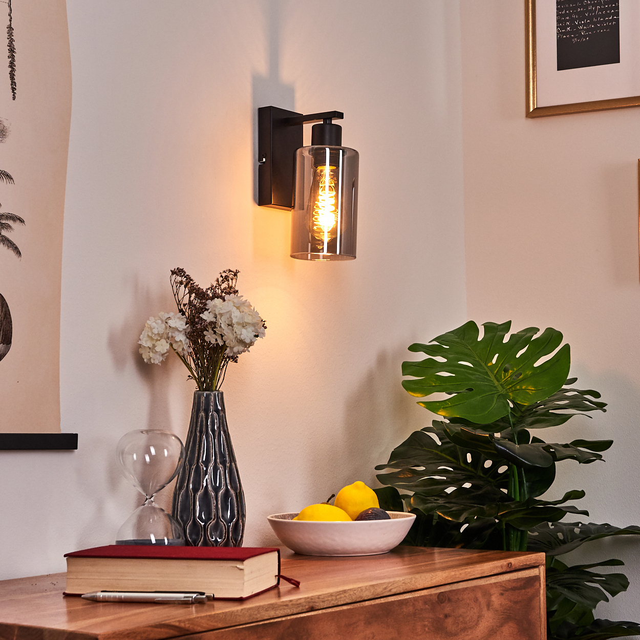 Eine Wandlampe beleuchtet einen Holztisch mit Vase, Blumen, Obst und einem Buch. Im Hintergrund eine Monstera-Pflanze.