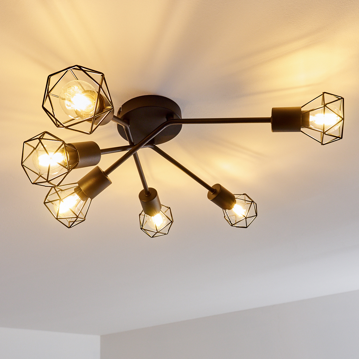 Eine schwarze Deckenleuchte mit sechs Glühbirnen in geometrischen Käfigen. Die Glühbirnen leuchten vor einer weißen Decke.