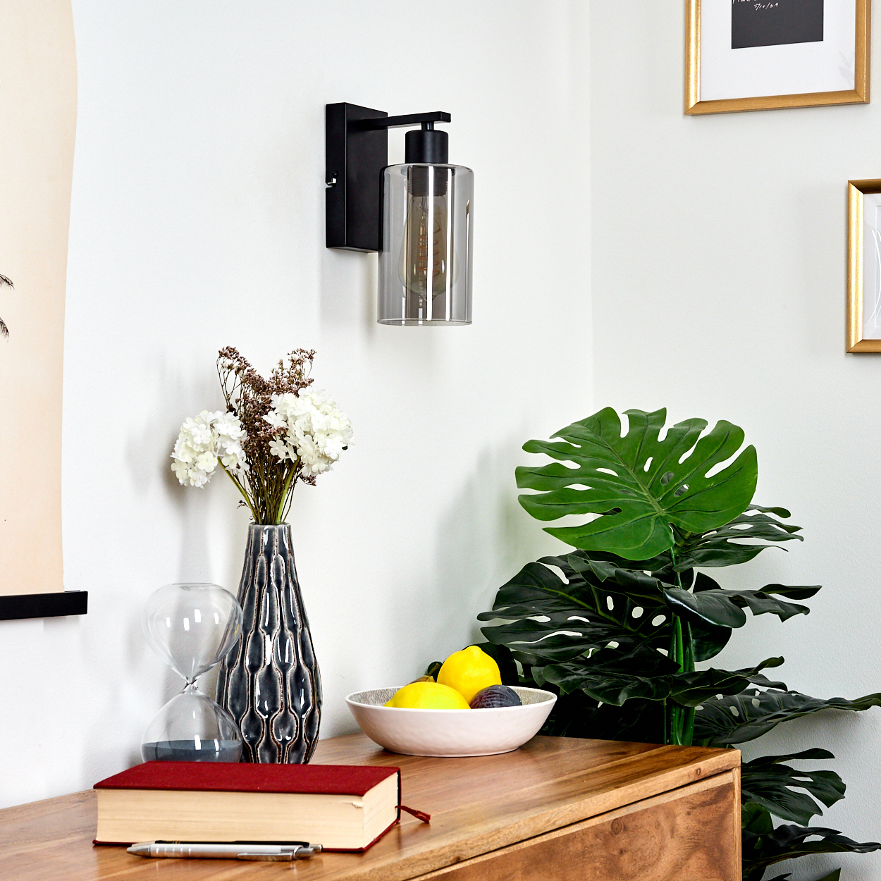 Eine Zimmerecke mit schwarzer Wandleuchte, Blumenvase, Obstschale und einem roten Buch auf einem Holztisch.