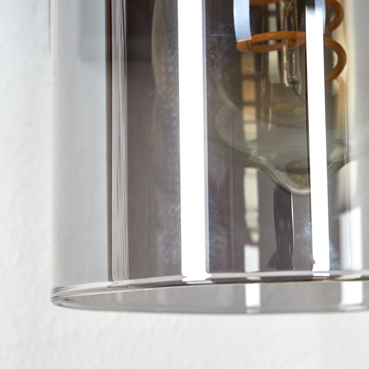 Nahaufnahme einer Leuchte. Ein klarer Glaszylinder umschließt eine Glühbirne mit sichtbarem orangefarbenem Glühfaden. Der Hintergrund ist eine weiße Wand.