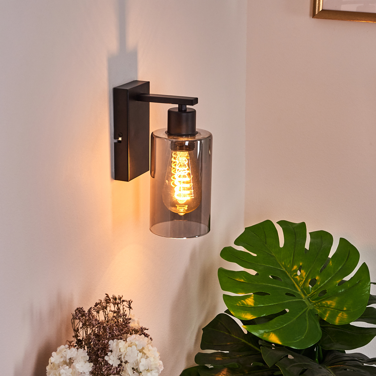 Eine Wandlampe mit schwarzem Rahmen und Glasschirm. Eine leuchtende Glühbirne im Inneren. Rechts eine grüne Pflanze, darunter Blumen.