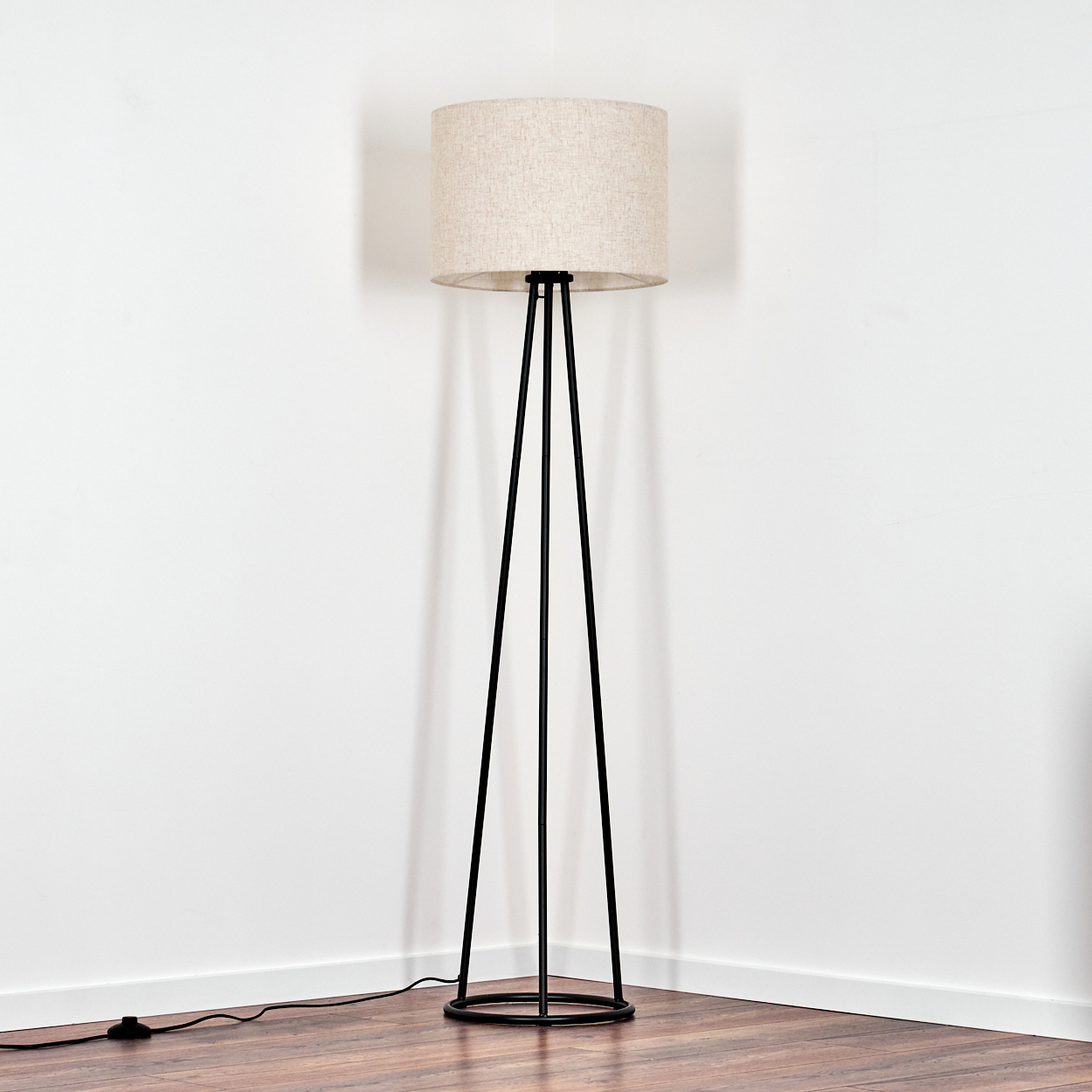 Eine Stehlampe mit beigem Lampenschirm und schwarzen Beinen steht in einer Ecke. Der Boden ist aus Holz, die Wände sind weiß.