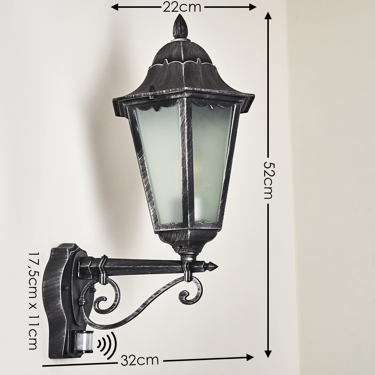 Eine an der Wand montierte, schwarze Außenlampe mit Milchglasscheiben. Die Lampe hat Zierschnörkel und einen Bewegungssensor. Abmessungen sind angegeben.