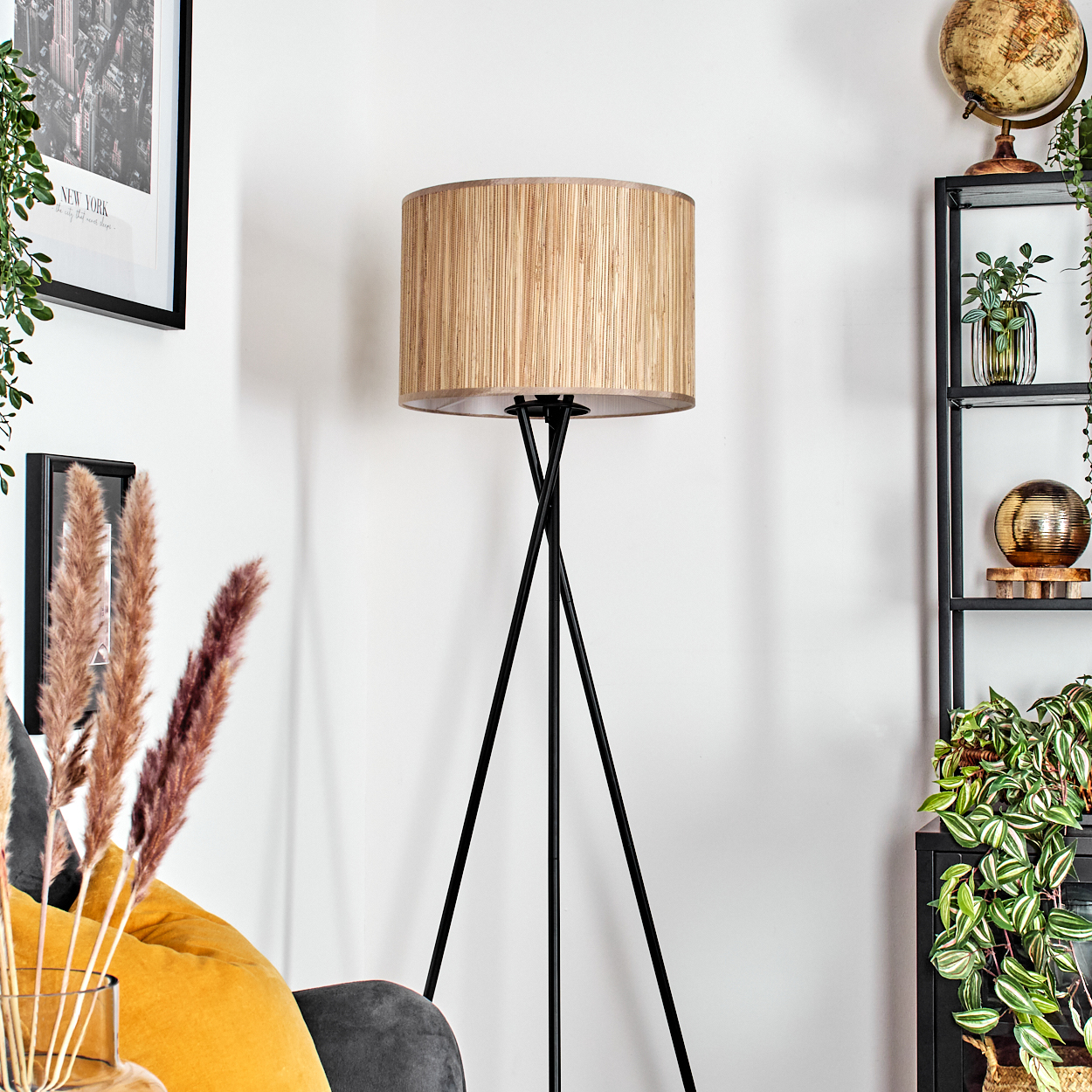 Eine Stehlampe mit beigem Lampenschirm steht auf einem schwarzen Stativ. Rechts ein Regal mit Pflanzen und Deko. Ein gerahmtes Foto hängt an der Wand.