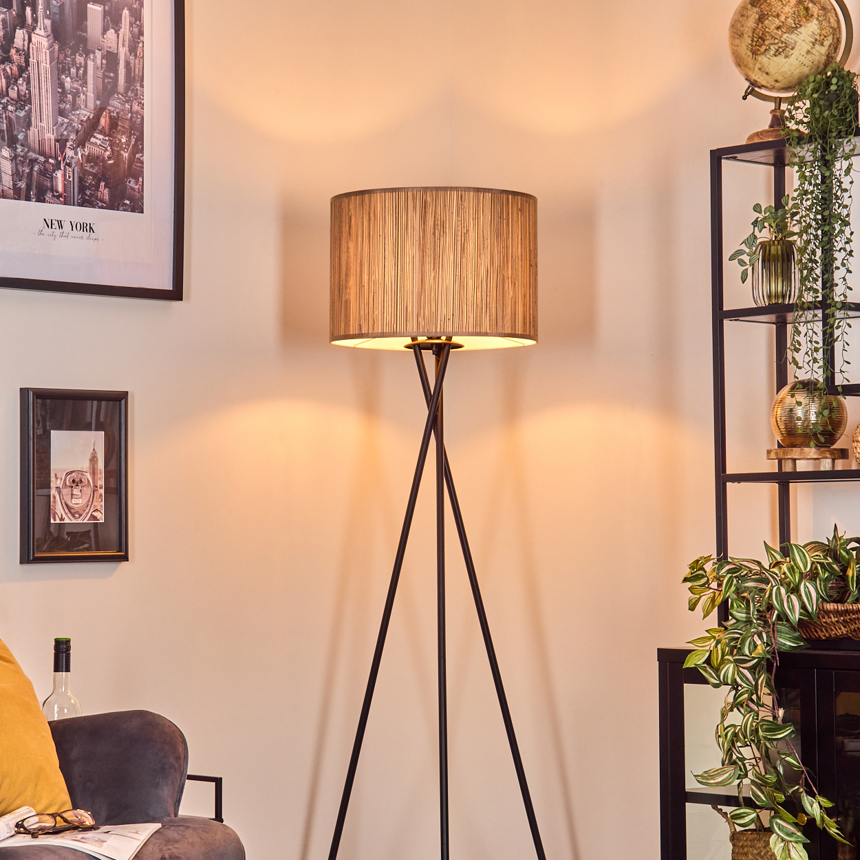 Eine Stehlampe mit strukturiertem Schirm steht in einem Raum. Ein New York City-Poster, ein Globus und Pflanzen sind ebenfalls sichtbar.