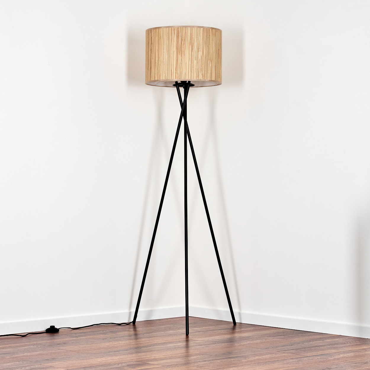 Eine Stehlampe mit zylindrischem, beigem Lampenschirm steht auf einem Holzboden. Sie hat schwarze Dreibeinbeine vor einer weißen Wand.