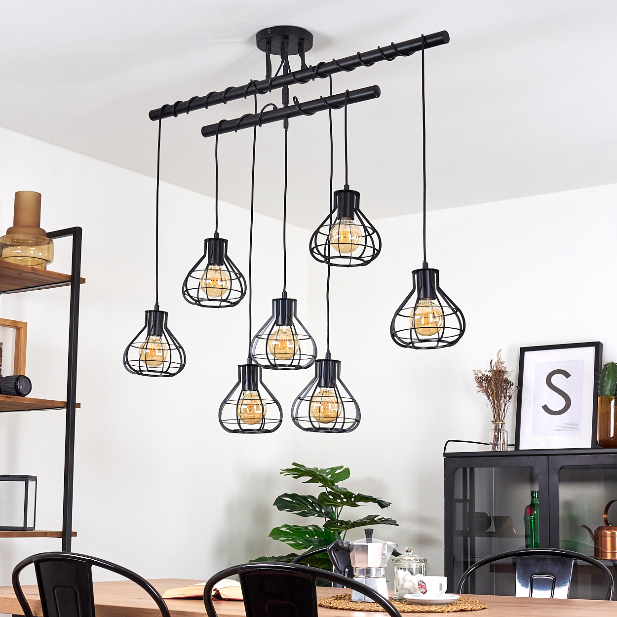 Schwarze Pendelleuchten mit Drahtkäfigen hängen über einem Holztisch. Ein schwarzes Metallregal, ein Schrank und eine Pflanze sind ebenfalls sichtbar.