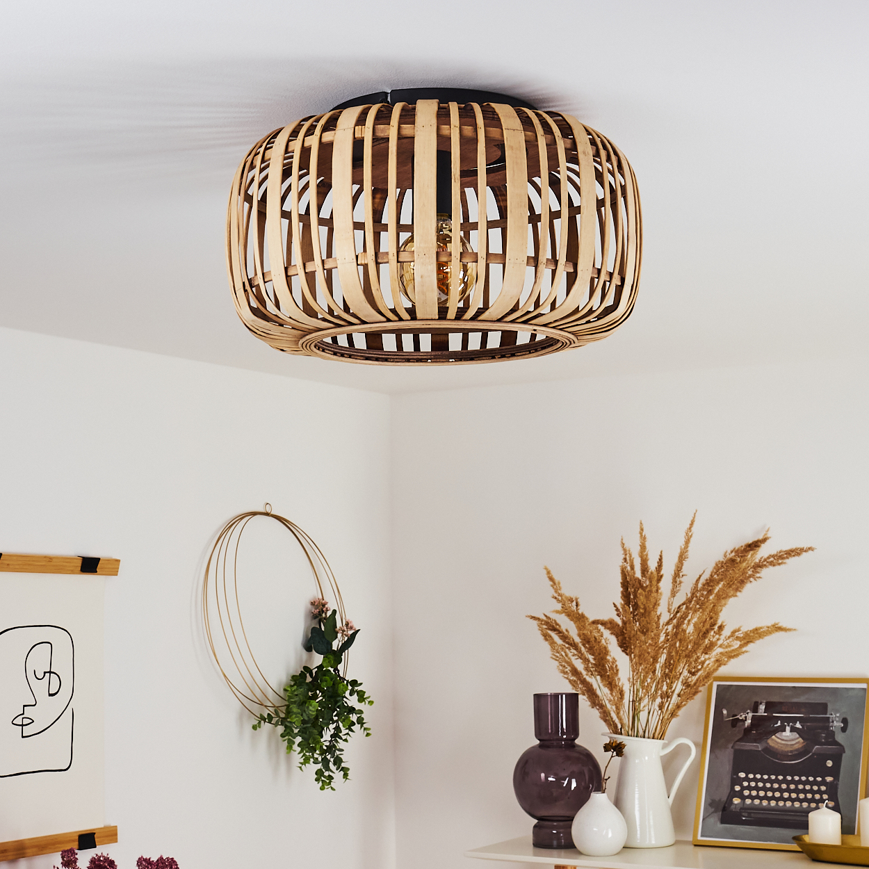 Eine Deckenlampe aus gewebtem Bambus hängt über einem weißen Raum mit Dekoration, darunter ein goldener Kranz mit Pflanzen und ein Bild.
