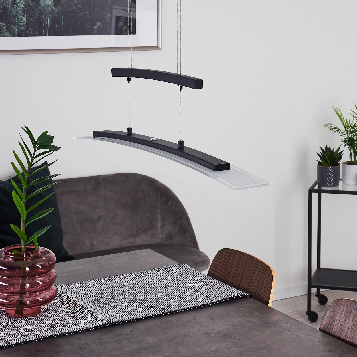 Ein modernes Esszimmer mit einer schwarzen und klaren Glas-Pendelleuchte. Ein Tisch mit gemustertem Läufer, einer Vase und einem grauen Samtstuhl.