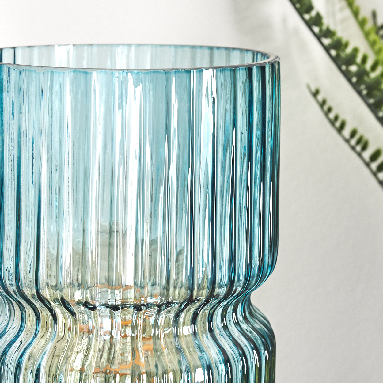 Eine geriffelte, blaue Glasvase. Im Hintergrund ist ein Farn. Die Vase hat einen ausgestellten, gebogenen Boden und eine gerade, zylindrische Oberseite.