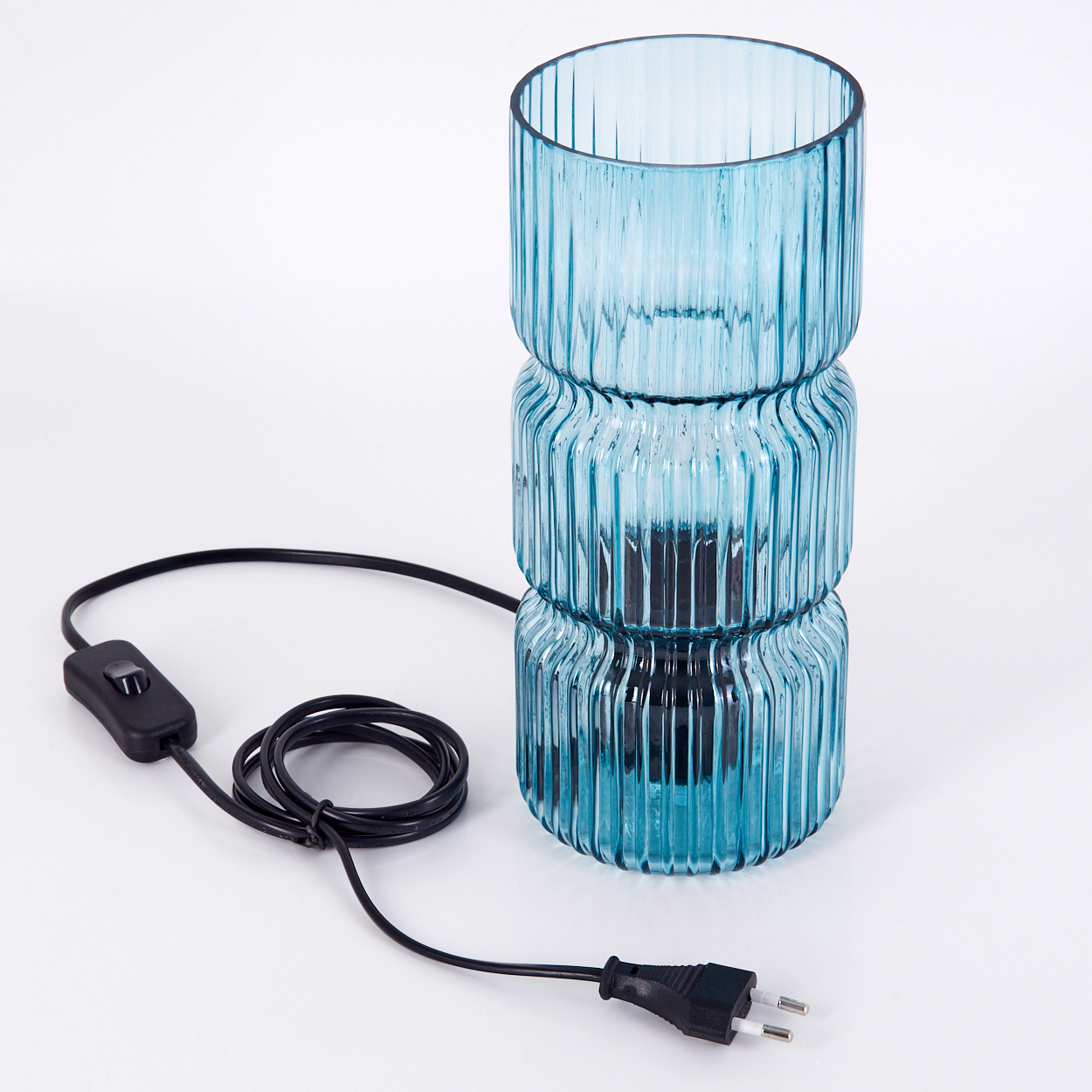 Eine blaue Glaslampe mit schwarzem Kabel und Stecker steht auf einer weißen Oberfläche. Die Lampe ist zylindrisch und hat eine geriffelte Textur.