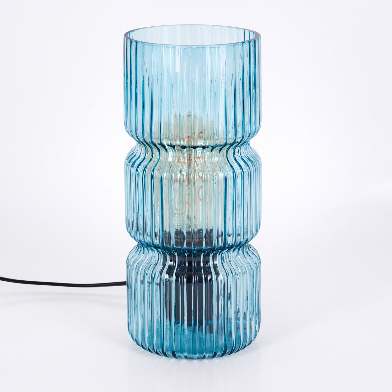 Eine blaue Glaslampe mit geripptem Design. Eine Glühbirne ist sichtbar. Ein schwarzes Netzkabel kommt aus dem Sockel. Weißer Hintergrund.