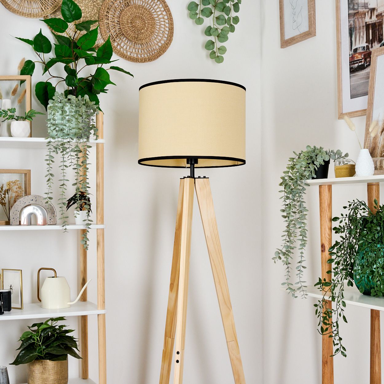Eine beige Lampe mit einem Stativ aus Holz. Regale mit Pflanzen, Bildern und Dekorationsartikeln sind sichtbar. Der Hintergrund ist weiß.