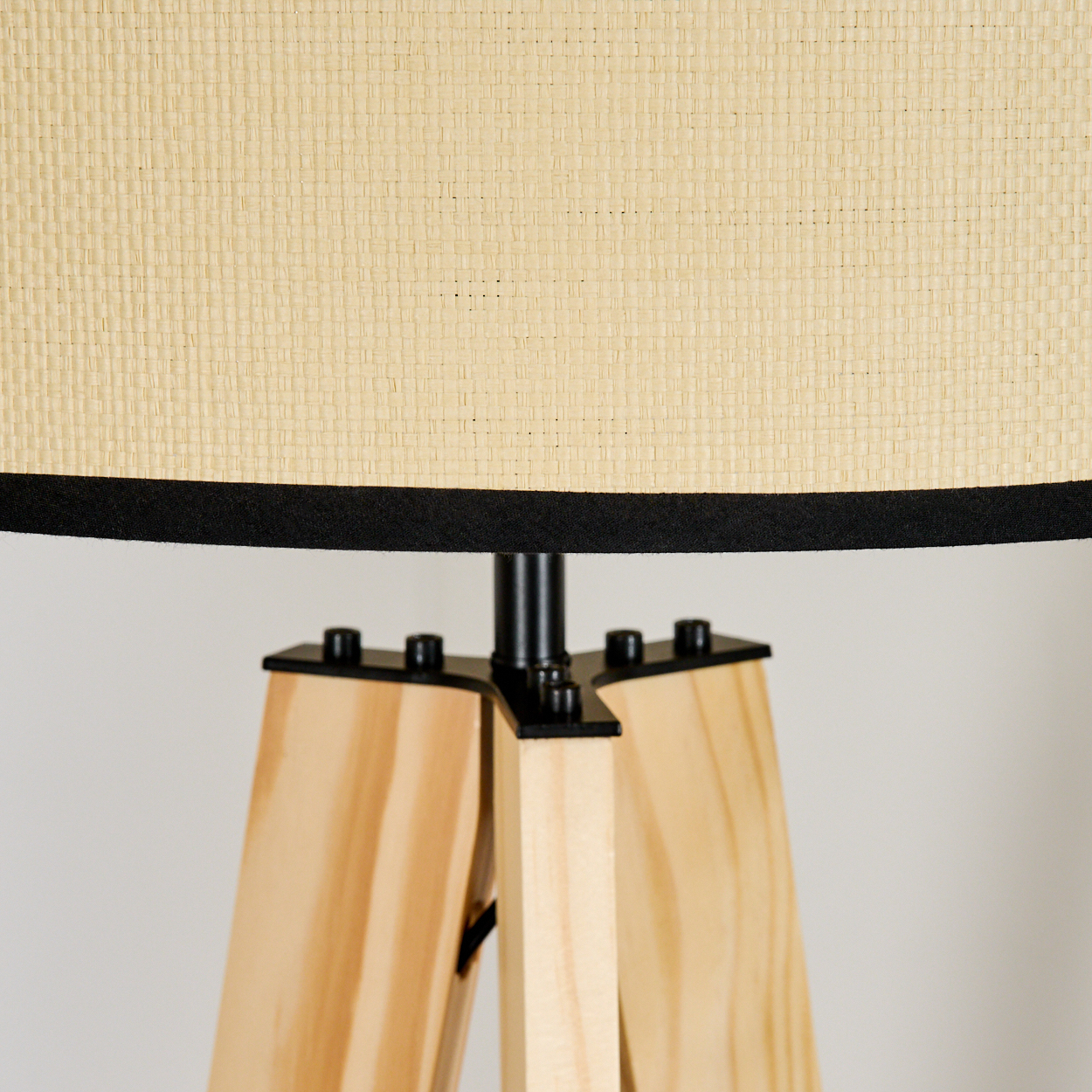 Nahaufnahme einer Lampe mit beigem Lampenschirm, schwarzem Rand und Holzbeinen. Der Hintergrund ist eine weiße Wand.