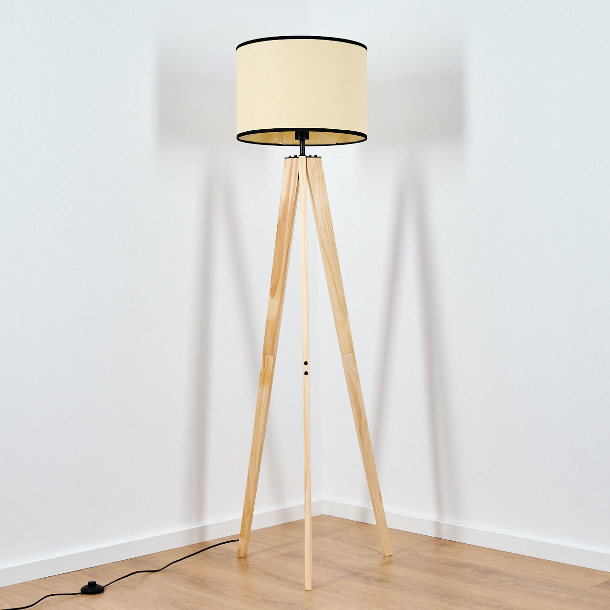 Eine Holz-Dreibein-Stehlampe mit beigem Lampenschirm steht in einer Ecke eines Raumes mit weißen Wänden und Holzboden.