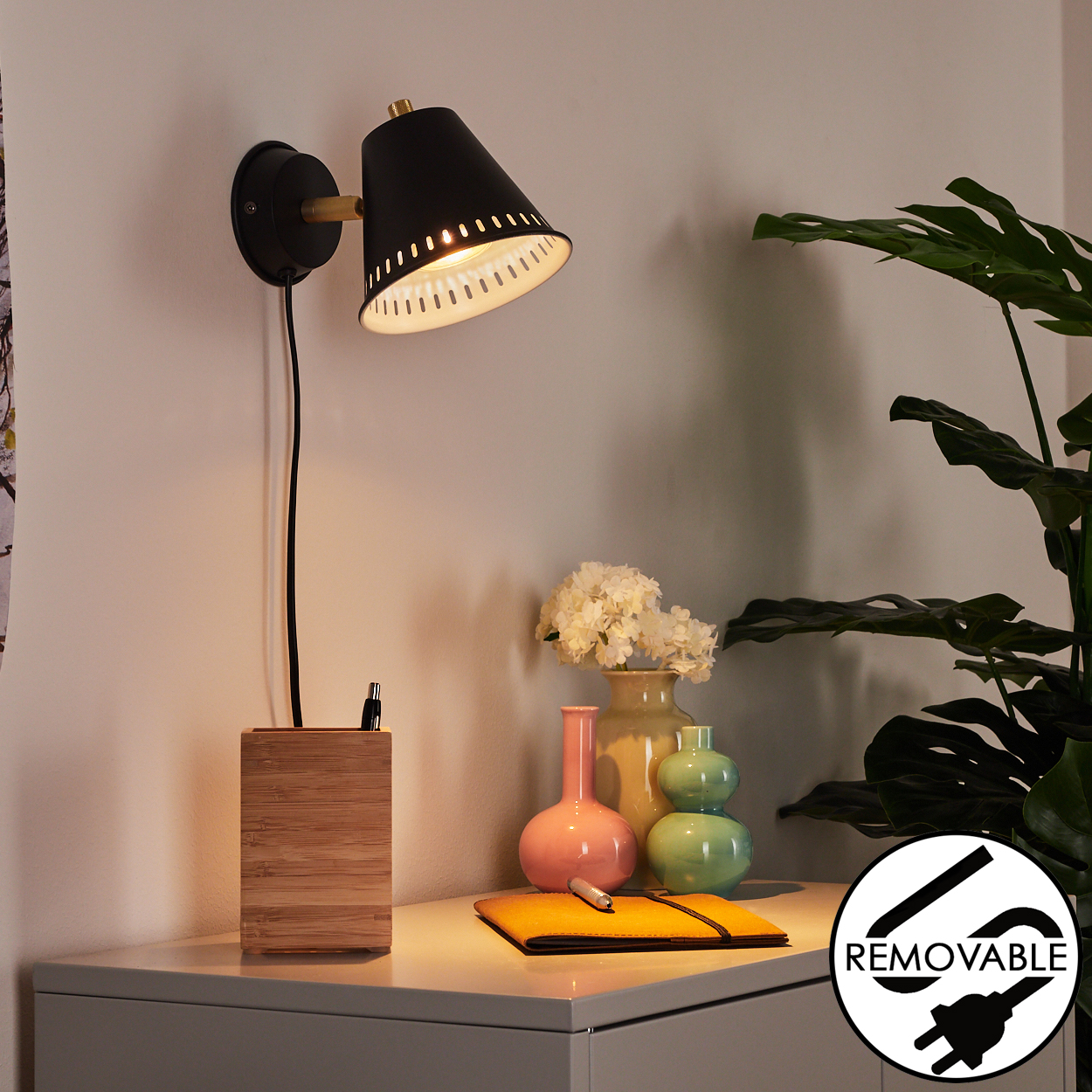 Eine schwarze Wandlampe mit Holzsockel beleuchtet einen Schreibtisch mit Vasen, einem Notizbuch und einer Pflanze im Hintergrund.