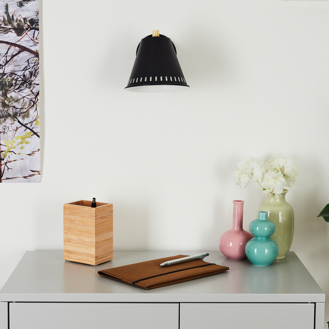 Eine schwarze Wandlampe, ein Schreibtisch mit einem hölzernen Stifthalter, einem braunen Notizbuch und einem Stift. Vasen mit Blumen stehen auf dem Schreibtisch.