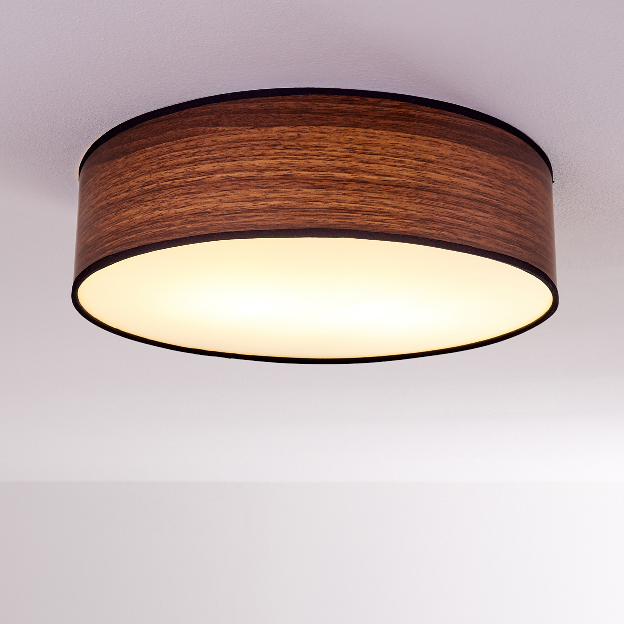 Gezeigt wird eine Deckenlampe mit Holzschirm. Die Lampe ist rund und strahlt warmes Licht aus. Der Hintergrund ist eine weiße Decke.