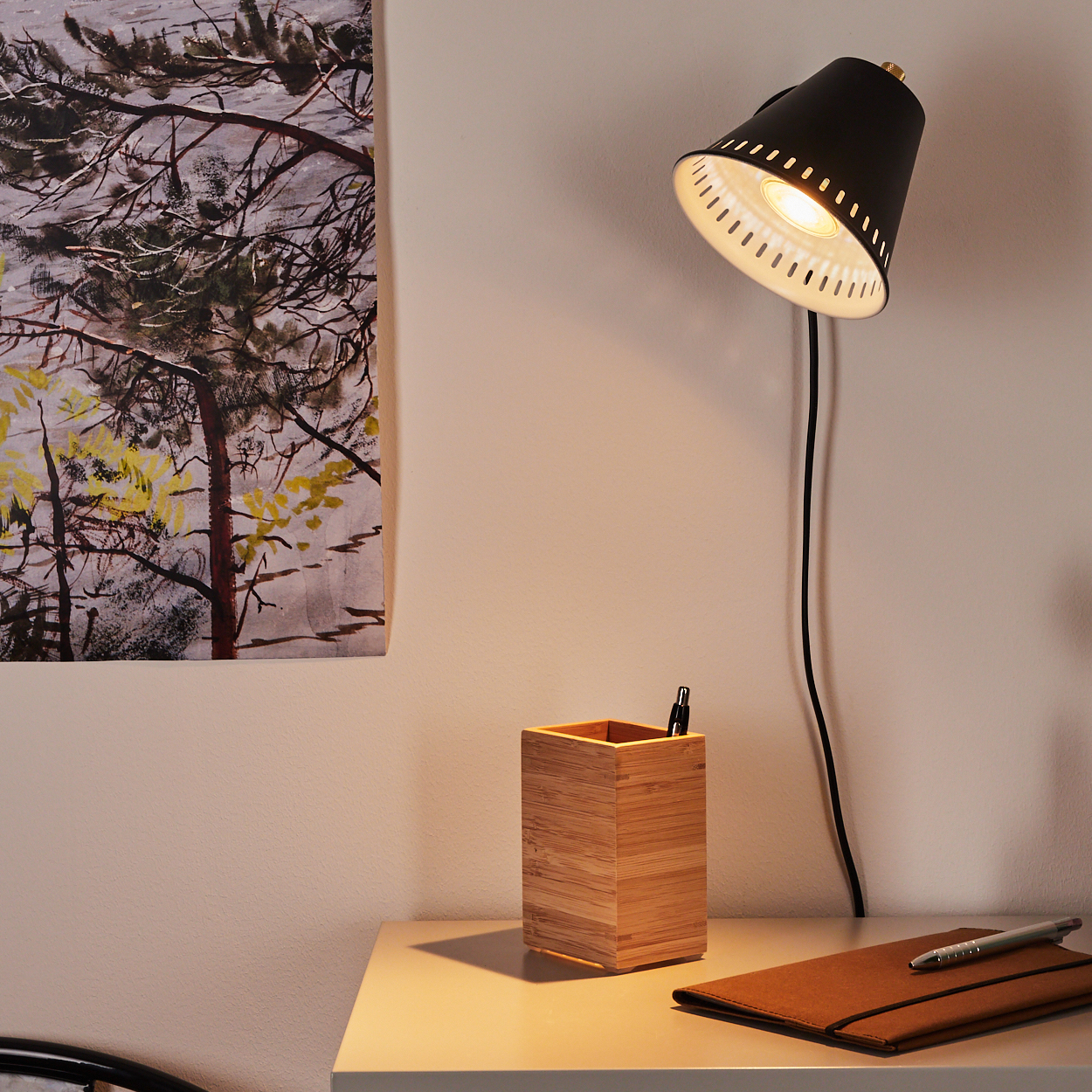 Ein Schreibtisch mit einer schwarzen Lampe, einem Stifthalter aus Holz, einem Notizbuch und einem Gemälde von Bäumen. Die Szene ist gut beleuchtet.