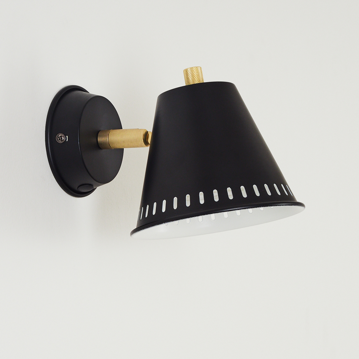 Eine schwarze Wandleuchte mit kegelförmigem Schirm, weißem Innenraum und Messingakzenten ist an einer weißen Wand befestigt.