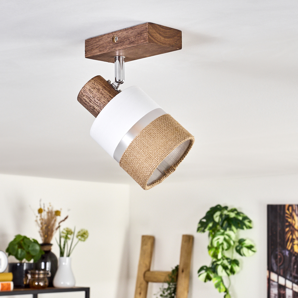 Eine Deckenlampe mit Holzsockel. Die Lampe hat einen weißen und Jute-Schirm. Eine Holzleiter und Pflanzen sind im Hintergrund.