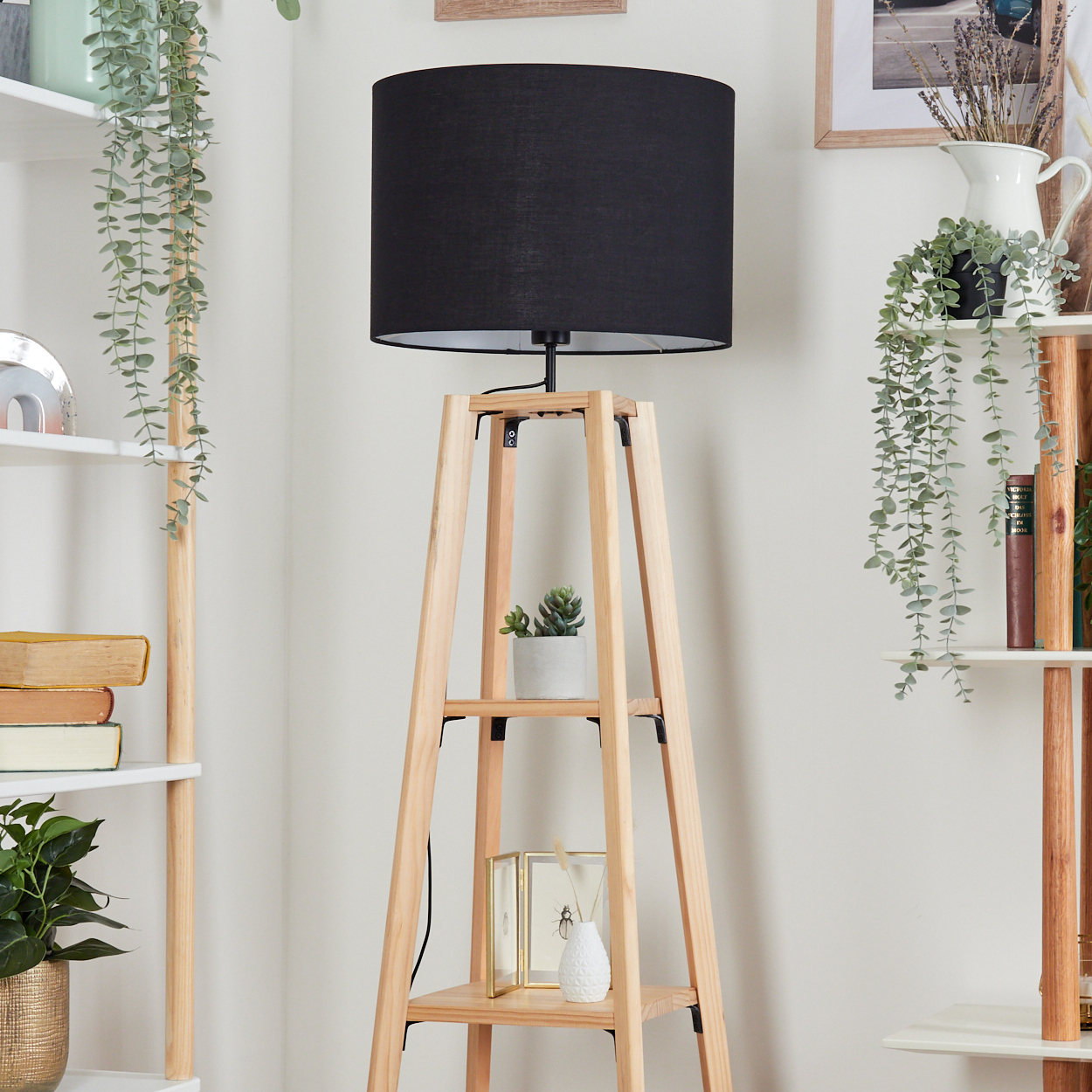 Eine Holzlampe mit schwarzem Lampenschirm steht vor einer weißen Wand. Regale mit Pflanzen und Büchern sind an den Seiten.