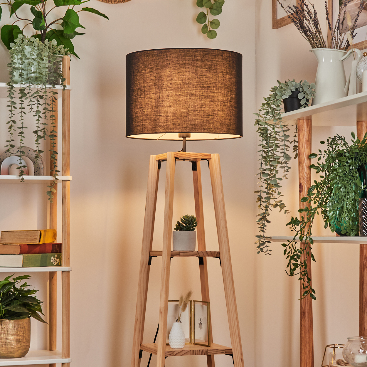 Eine hölzerne Stehlampe mit schwarzem Schirm. Regale mit Pflanzen und Büchern befinden sich auf beiden Seiten. Der Hintergrund ist eine helle Wand.