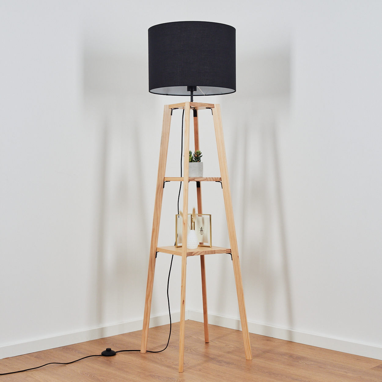 Eine Stehlampe mit schwarzem Lampenschirm auf Holzgestell. Regale mit Pflanze, Bilderrahmen. Hintergrund ist eine weiße Wand.