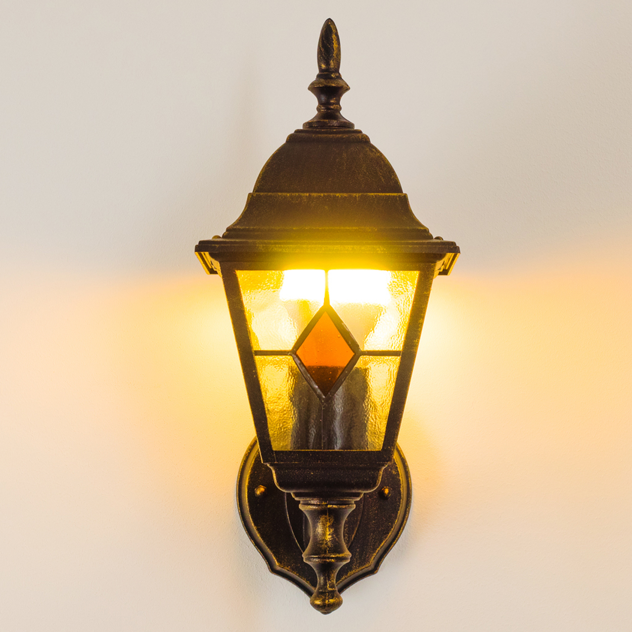 Eine an der Wand befestigte Außenlampe mit dekorativem Design. Sie hat eine bronzefarbene Oberfläche und strahlt warmes, gelbes Licht aus.