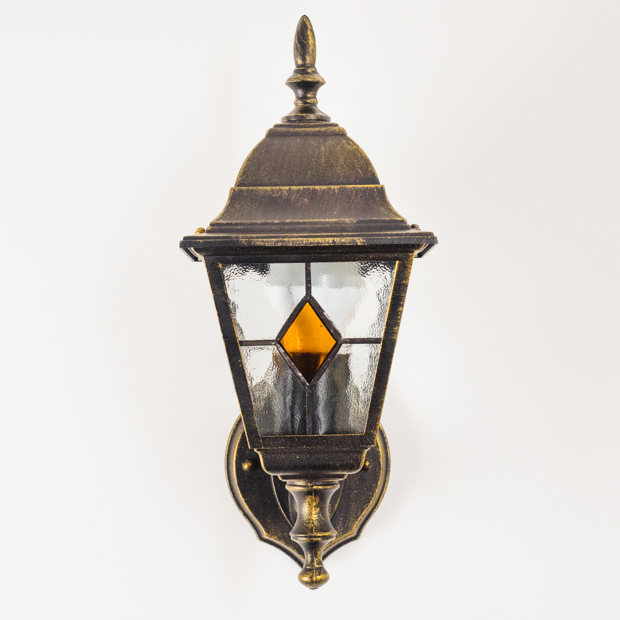 Eine verzierte, bronzefarbene Außenlampe mit Glasscheiben und einem dekorativen Zierrat, montiert an einer weißen Wand.