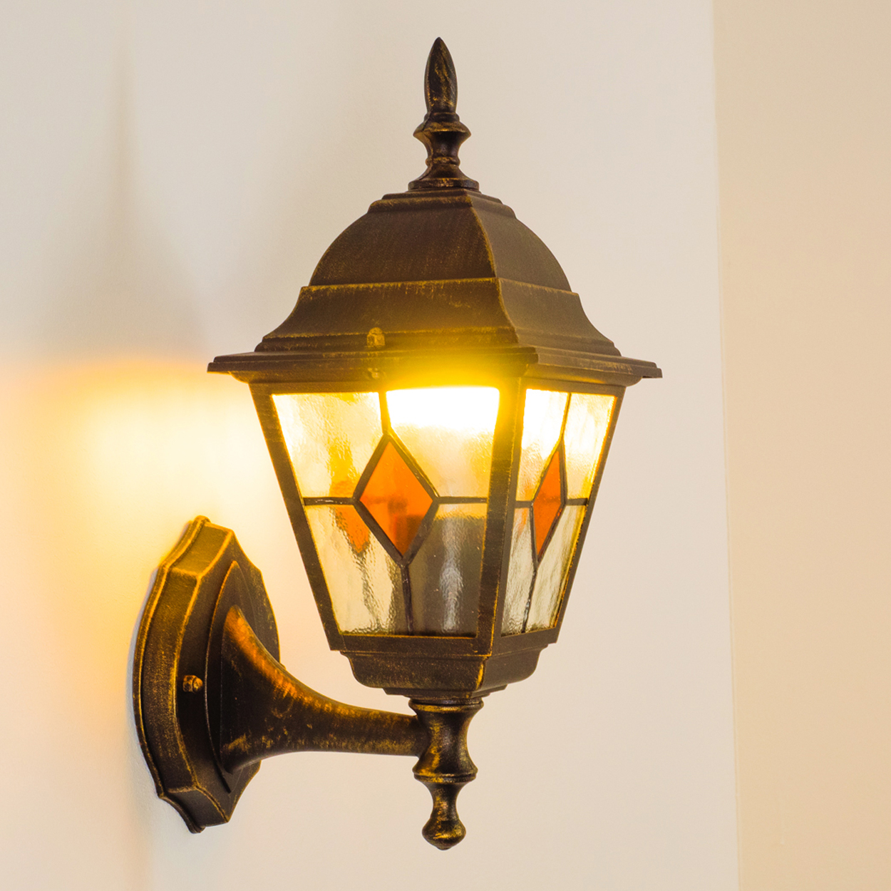 Eine verzierte bronzefarbene Wandlampe mit Glasmalerei. Die Lampe ist beleuchtet und wirft einen warmen Schein auf eine weiße Wand.